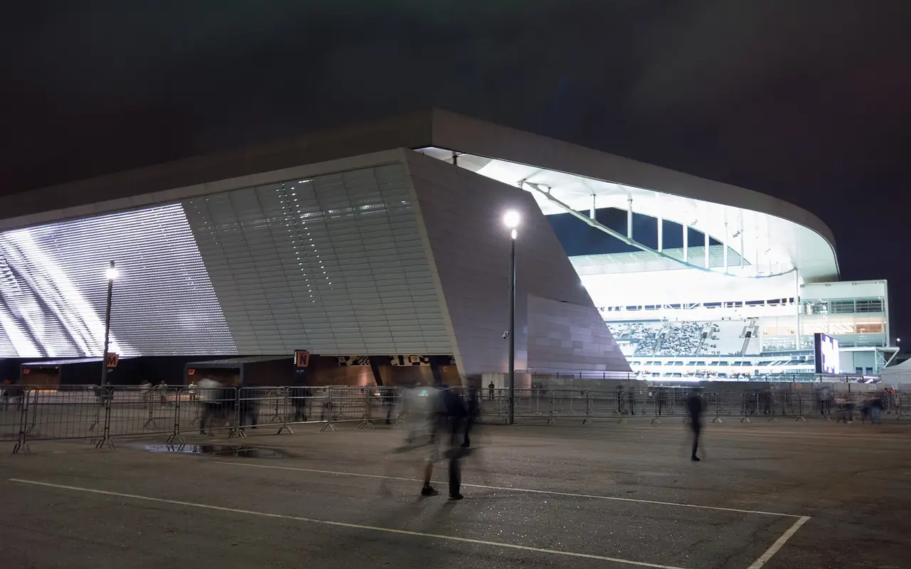 Corinthians Arena in São Paulo at night: a wide open asphalt apron in front of the stadium, with crowd-control barriers and people dispersing across open ground.