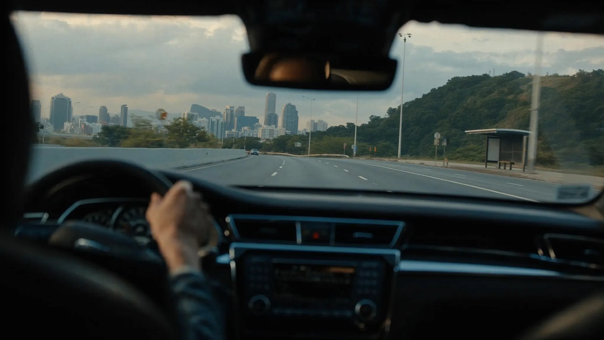 Rear-seat view from a private executive sedan moving along an open Rio de Janeiro expressway at first light, the city skyline soft in the background and an empty bus stop at the roadside.